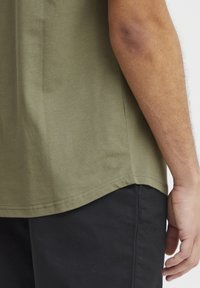 Olive green cotton t-shirt with a rounded hem and short sleeves, paired with black pants. The fabric is smooth with a slight sheen.