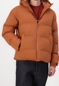 Hnědá prošívaná bunda s předním zipem a kapucí, s hladkou texturou a mírně volným střihem, doplněná tmavými džínami.