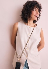 Bēša lina veste bez piedurknēm ar pieciem priekšējiem pogām un vieglu A veida formu. Ir ķēdes siksniņa ar tālruņa maciņa turētāju. Gaišs fons.