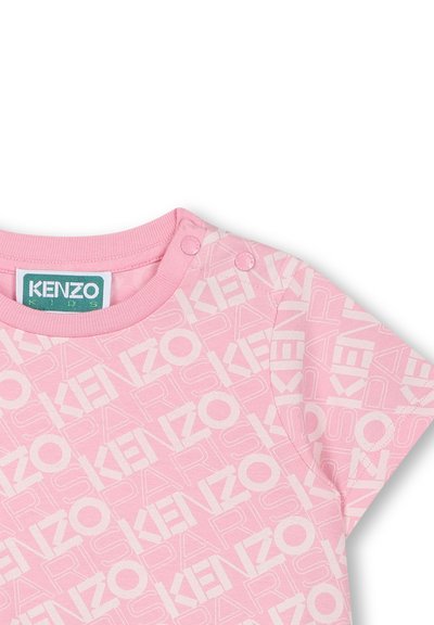 Różowa bawełniana koszulka z krótkim rękawem, z powtarzającym się białym wzorem logo. Zapięcia na napy przy ramieniu ułatwiają zakładanie.