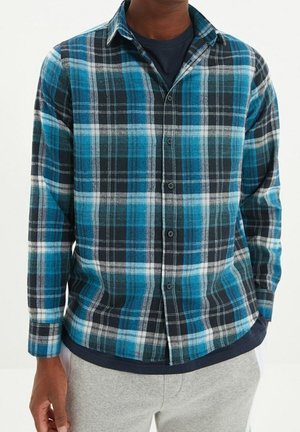 Uomo che indossa una camicia a quadri blu, nera e bianca sopra una maglietta scura, con pantaloni grigio chiaro, mani rilassate ai lati.