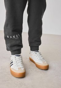 Białe sneakersy z gumowymi podeszwami, z granatowymi i białymi paskami, w połączeniu z ciemnoszarymi spodniami dresowymi z logiem "BAKER" blisko dołu.