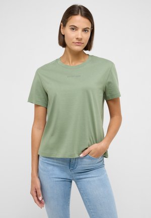 Frau mit kurzen braunen Haaren, die ein hellgrünes Kurzarmshirt und hellblaue Jeans trägt, posiert mit einer Hand in der Hosentasche.