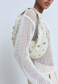 Sac à main de couleur crème en matériau lisse, orné d'accents sphériques métalliques. Porté par-dessus un pull en tricot blanc à mailles ouvertes.