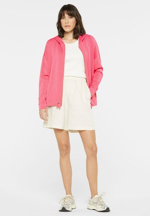Rosa Zip-Up Hoodie mit lockerer Passform, ausgestattet mit Kapuze und Kordelzug, kombiniert mit einem weißen T-Shirt und beigen Shorts, abgerundet mit Sneakers.