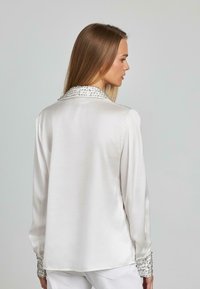 Camicia in satin bianco con colletto e polsini decorati con perline. Maniche lunghe con una vestibilità leggermente ampia. Tessuto liscio.