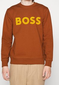 Sudadera marrón con un gran logo amarillo "BOSS" en el pecho. Cuenta con puños y dobladillo de canalé, fabricada con un tejido suave.
