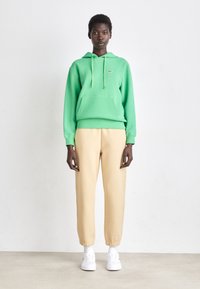 Felpa verde con tasca frontale e cordino, abbinata a pantaloni della tuta beige a gamba stretta e sneaker bianche. Design semplice e informale.