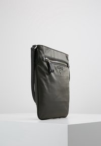 Sort lædertaske med crossbody-strop, glat tekstur, frontlomme med lynlås og logodetalje. Har en justerbar rem for bekvemmelighed.