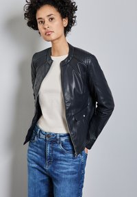 Veste bomber en cuir noir avec des détails matelassés aux épaules, associée à une chemise beige clair et un jean bleu. Texture lisse et design ajusté.