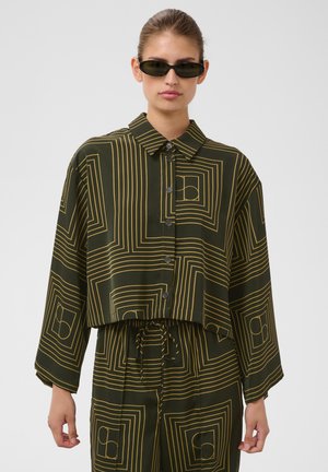 Femme portant une chemise et un pantalon assortis vert foncé avec des motifs géométriques carrés jaunes et des lunettes de soleil noires étroites, sur un fond uni.