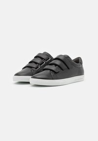 Baskets en cuir noir avec deux larges brides en Velcro, un bout pointu et une semelle en caoutchouc blanc. Texture lisse avec des détails de couture subtils.