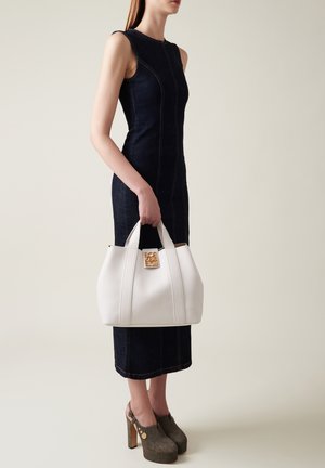 Borsa a tracolla bianca in pelle con finiture dorate, forma arrotondata, cuciture verticali, tenuta da una persona che indossa un vestito attillato in denim scuro e scarpe con plateau.