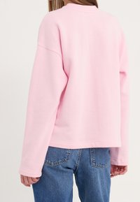 Sudadera rosa, de gran tamaño, con cuello redondo, mangas largas y una textura suave. Combinada con jeans de mezclilla azules, mostrando un ajuste casual.
