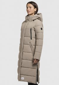 Lang beige pufferfrakke med hætte, featuring quiltet tekstur, sorte lynlåslommer og et logo patch nær kanten. Glat materiale.