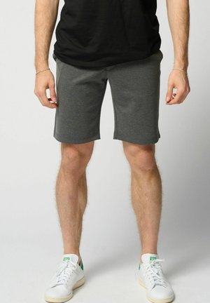 Mann trägt dunkelgraue Shorts, schwarzes T-Shirt und weiße Sneaker und steht vor einem schlichten hellen Hintergrund.