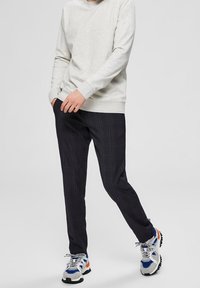 Šedá mikina, tmavé kárované kalhoty a sportovní boty s vícebarevným provedením. Mikina má kulatý výstřih; kalhoty jsou úzkého střihu a hladké textury.