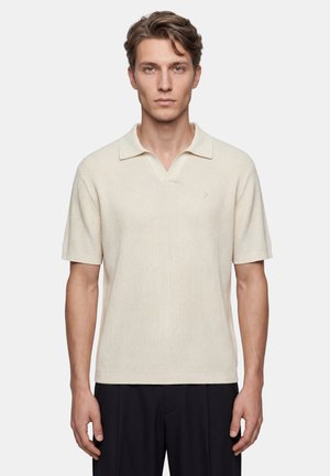 Giovane uomo con capelli castano chiaro che indossa una polo a maniche corte color crema in maglia e pantaloni neri, in piedi davanti a uno sfondo bianco uniforme.