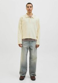 Pull jaune clair avec un col et trois boutons, associé à un jean baggy bleu clair et des chaussures marron, se tenant devant un fond uni.