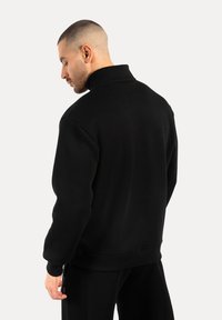 Muž s krátkými vlasy, oblečený v černé mikině s vysokým límcem a černých kalhotách, stojící bokem na světlém jednobarevném pozadí.