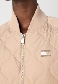 Gros plan d'une veste matelassée beige avec une fermeture éclair argentée et un écusson logo Tommy Jeans sur la poitrine, portée par une personne à la peau foncée.