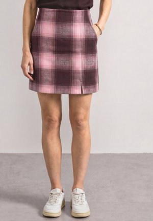 Personne portant une jupe à carreaux rose et violette avec une fente devant, des baskets blanches et un haut noir, debout sur un sol gris.