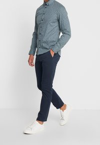 Hombre con una camisa de botón de flores azules, pantalones ajustados navy y zapatillas blancas caminando con una mano en el bolsillo contra un fondo blanco.