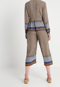Mönstrad jumpsuit i bruna, blåa och orange färger; har långt ärmar, elastisk midja, korta vida ben och geometrisk design.
