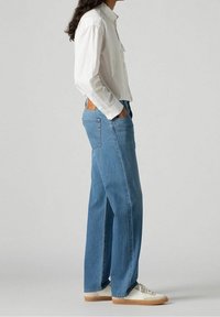 Jeans de mezclilla azul con un corte recto, lavado claro y un ligero desvanecimiento. Combinados con una camisa blanca de botones y zapatillas blancas.