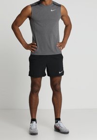 Szary bezrękawnik sportowy z teksturowanej tkaniny, w zestawieniu z czarnymi szortami. Posiada białe logo Nike. Noszony z szaro-czarnymi butami sportowymi.