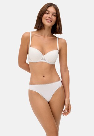 Mujer con cabello castaño hasta los hombros sonriente, vistiendo un sujetador de encaje blanco y pantis blancos a juego, de pie con las manos detrás de la espalda.