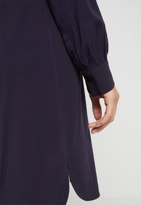 Mörkblå skjorta med långa, puffiga ärmar och knäppte manschetter. Tyget är slätt med en lätt lyster, och nederkanten har sidoinsnitt.