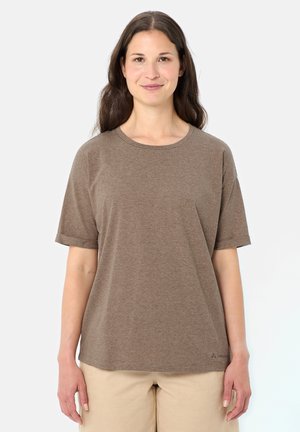 Frau mit langen braunen Haaren, die ein braun gestreiftes Kurzarmhemd und beige Hosen trägt, vor weißem Hintergrund stehend.