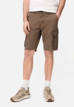 Mann trägt braune kniehohe Cargo-Shorts mit Klappen- und Reißverschlusstaschen, beige Sneaker, weiße Socken und ein in die Hose gestecktes weißes Hemd, steht.