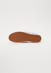 Semelle en caoutchouc avec un motif brun texturé, bordée d'une semelle intermédiaire blanche, présentant un dessus noir visible depuis le talon.