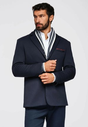 Marineblazer mit tailliertem Schnitt, präsentiert mit einer roten Akzenttasche, über einem weißen Hemd, kombiniert mit einem gestreiften Schal und blauen Hosen.