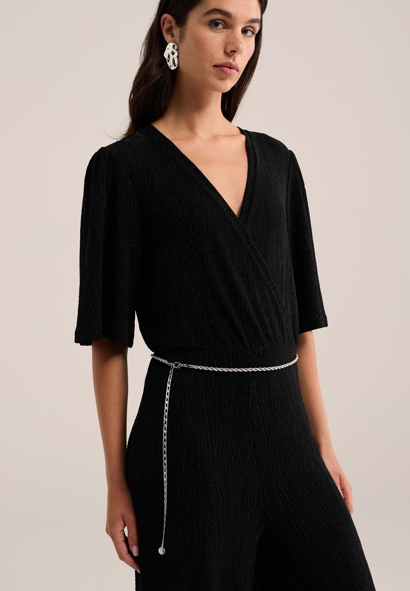 Femme portant une robe noire texturée à col en V avec des manches au coude, une fine ceinture chaîne argentée et de grandes boucles d'oreilles blanches abstraites.