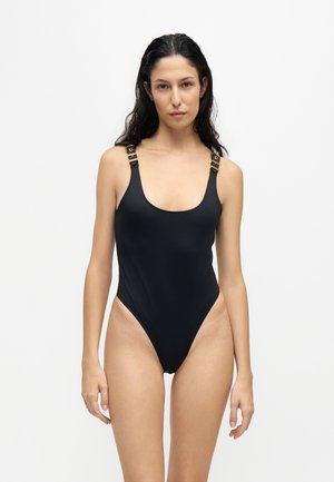 Femme aux longs cheveux foncés portant un maillot de bain une pièce noir avec des bretelles boucle dorée, debout devant un fond blanc uni.