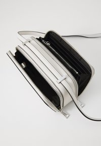 Bílá kožená crossbody taška se stříbrnými zipy, která má několik přihrádek a černý interiér, doplněná elegantním popruhem.