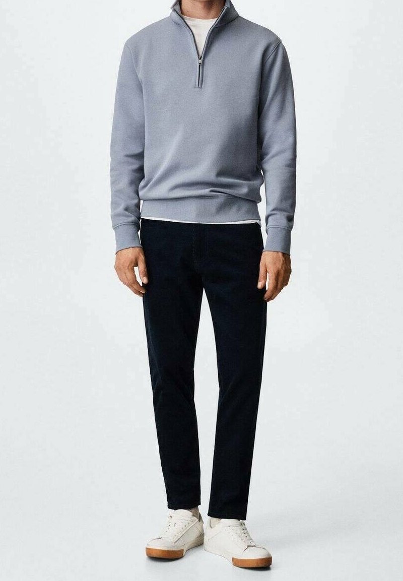 Felpa grigia con zip, pantaloni neri e sneakers bianche. La felpa ha un col alto e polsini a coste. Il tessuto sembra morbido e liscio.