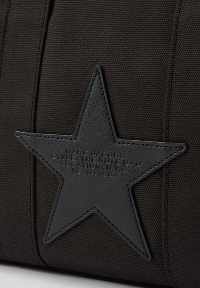 Svart kanvas totebag med et teksturert svart lærstjerne-patch som har preget merkevare detaljer. Sydd aksenter langs kantene.