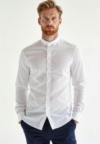 Biała koszula zapinana na guziki z kołnierzykiem stojącym, długim rękawem, subtelną teksturą i dopasowanym krojem. W zestawie z ciemnymi spodniami.