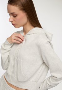 Felpa grigia chiara con zip, dal texture morbido, con tasche frontali e design aderente. Presenta un cappuccio e dettagli della cerniera in vista.
