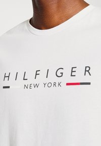 Detailní záběr osoby oblečené v bílém tričku s nápisem "HILFIGER NEW YORK" a černými, bílými a červenými grafickými prvky na hrudi.