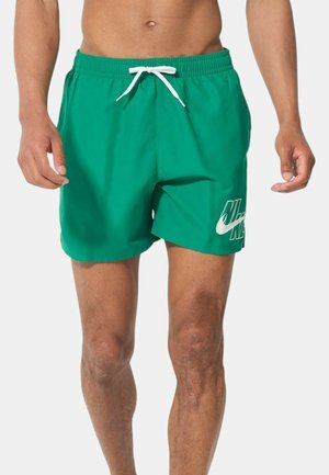 Uomo con pantaloncini da bagno verdi Nike con coulisse bianca e logo, in piedi su uno sfondo bianco uniforme.