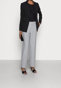 Sort blazer over en sort mønstret top, parret med lysegrå brede bukser og sorte åbne hæle; slankt, skræddersyet design.