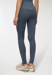 Granatowe legginsy wykonane z gładkiego, elastycznego materiału z wysokim pasem i detalem logo z tyłu, zestawione z beżowymi sneakersami.