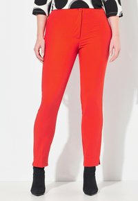 Červené slim-fit kalhoty vyrobené z elastického materiálu, s předním zapínáním na knoflík a nohavicemi po kotníky, v kombinaci s černými kotníkovými botami.