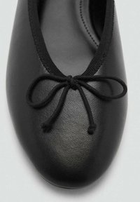 Bailarina de piel negra con punta redondeada, que presenta un detalle de lazo delgado en color negro en la parte frontal y una plantilla flexible y acolchada.