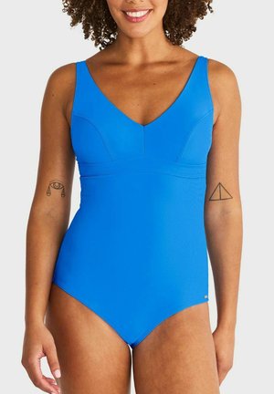 Lachende vrouw die een felblauw badpak draagt en tatoeages op beide bovenarmen laat zien tegen een effen achtergrond.